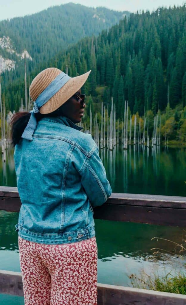 Woman wearing a sun hat and denim jacket overlooking the submerged forest at Kaindy Lake in Kazakhstan.