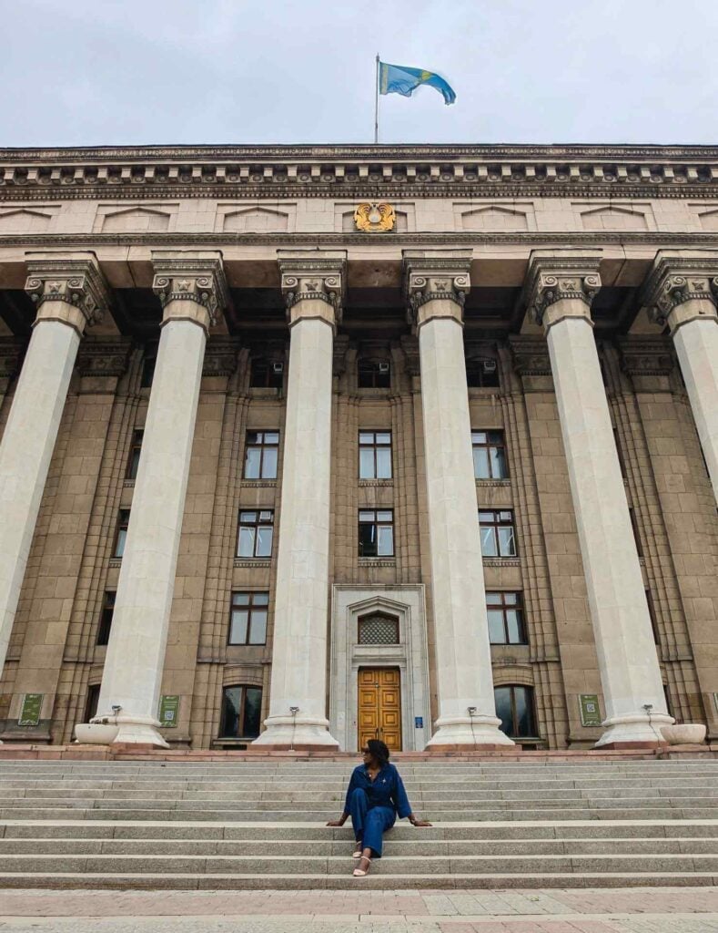Woman seated on the steps of the Kazakh-British Technical University beneath its grand columns and Kazakhstan flag in Almaty.