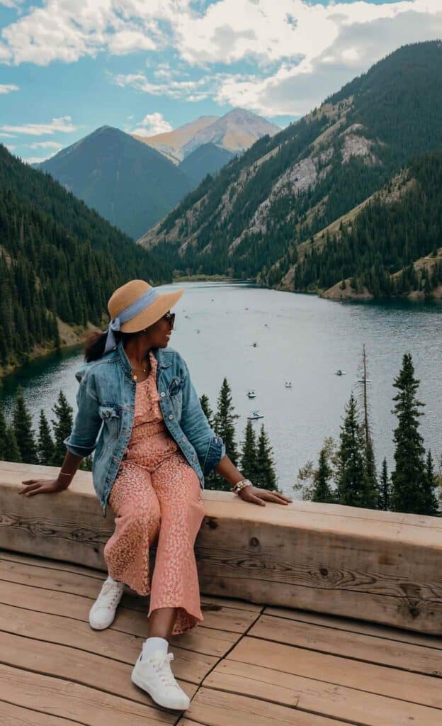Woman seated at a scenic viewpoint overlooking Lower Kolsai Lake, evergreen forests and mountains in Kazakhstan.