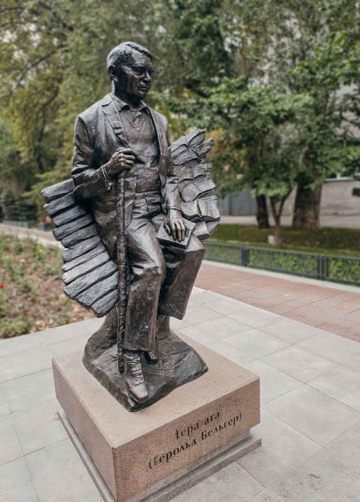 Bronze monument to writer Gerold Belger seated with a book and walking cane in a leafy public space in Almaty, Kazakhstan.