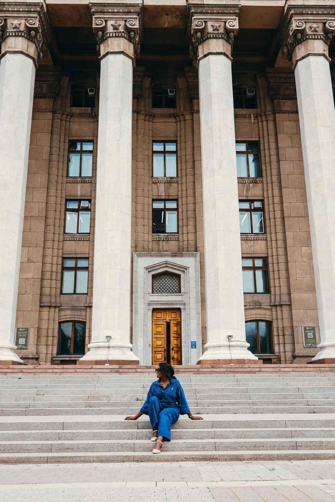 Black woman seated on the steps of the columned Kazakh-British Technical University building in Almaty, Kazakhstan.