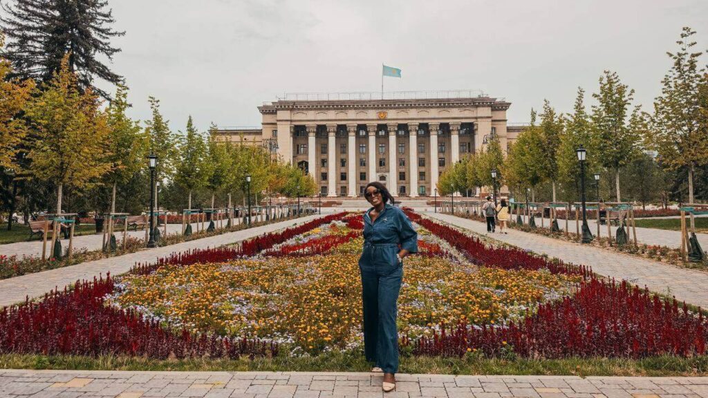 Black woman traveler standing among colorful flower beds in Astana Square, with the historic Kazakh-British Technical University building and Kazakhstan flag behind her in Almaty.