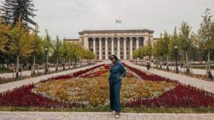 Black woman traveler standing among colorful flower beds in Astana Square, with the historic Kazakh-British Technical University building and Kazakhstan flag behind her in Almaty.