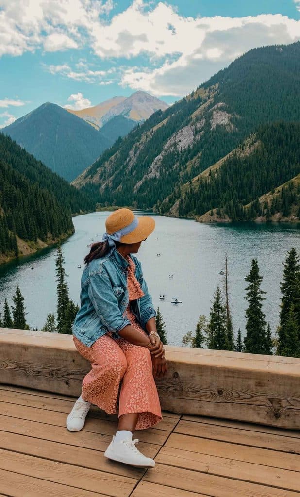 Black woman traveler overlooking Kolsai Lake in Kazakhstan, surrounded by forested slopes and the scenic Tian Shan Mountains.