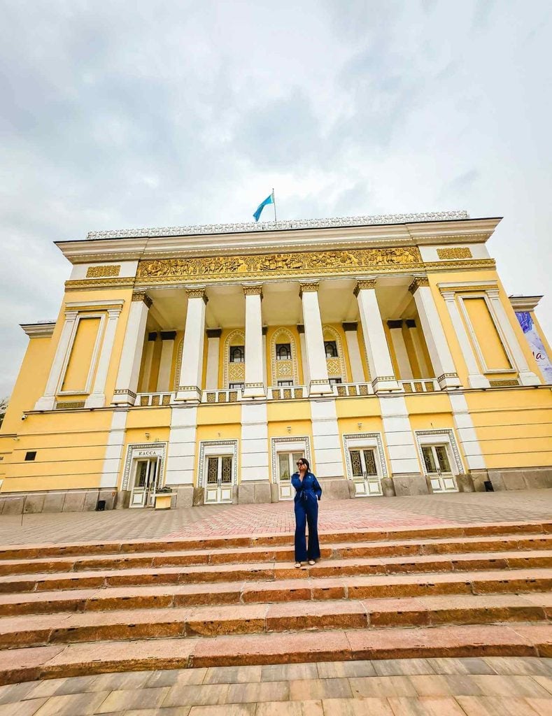Woman standing on the steps beside the yellow and white exterior of the Abai Opera House in Almaty, Kazakhstan.