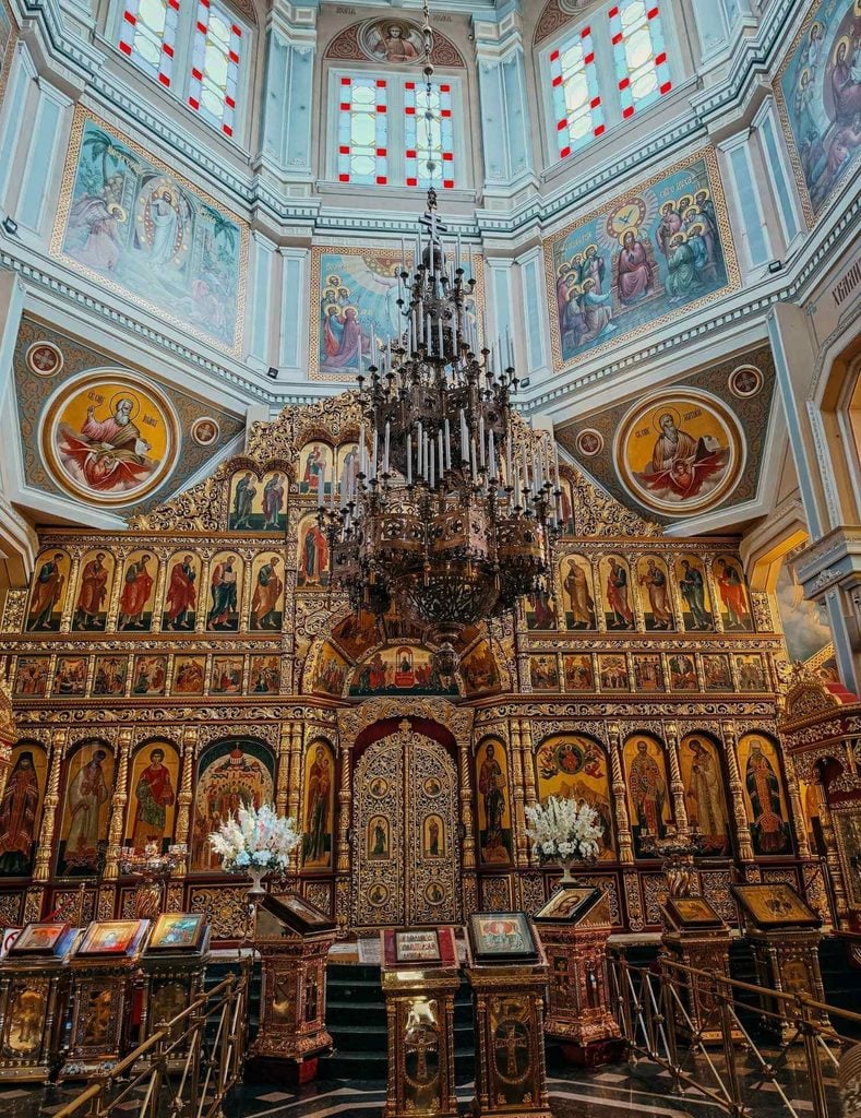 Ornate interior of Ascension Cathedral in Almaty featuring religious icons, murals, stained glass, and a grand chandelier.