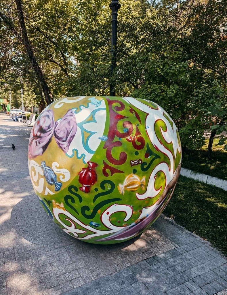 Colorful apple sculpture decorated with Kazakh patterns and candy illustrations on a tree-lined street in Almaty.
