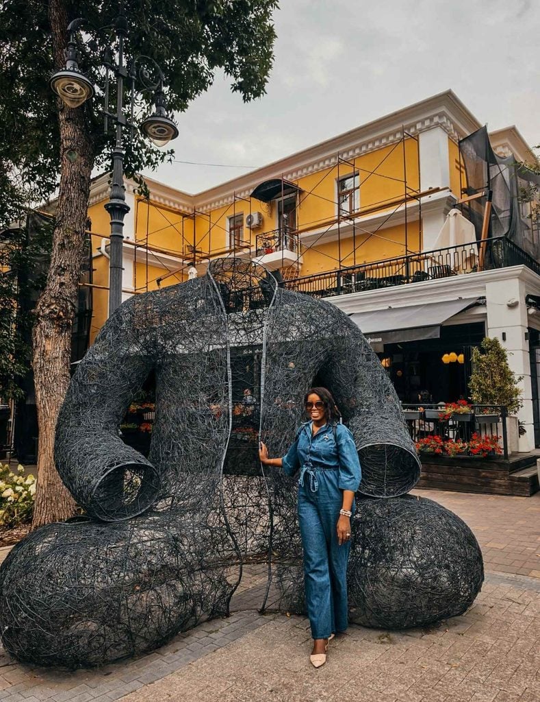 Woman standing beside the Kara Shapan metal art installation near Panfilov Street in Almaty, Kazakhstan.